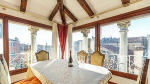 Une salle à manger avec une table dressée pour quatre personnes, une bouteille de vin et quatre verres. La pièce est dotée de poutres en bois au plafond et de grandes fenêtres offrant une vue sur les bâtiments.