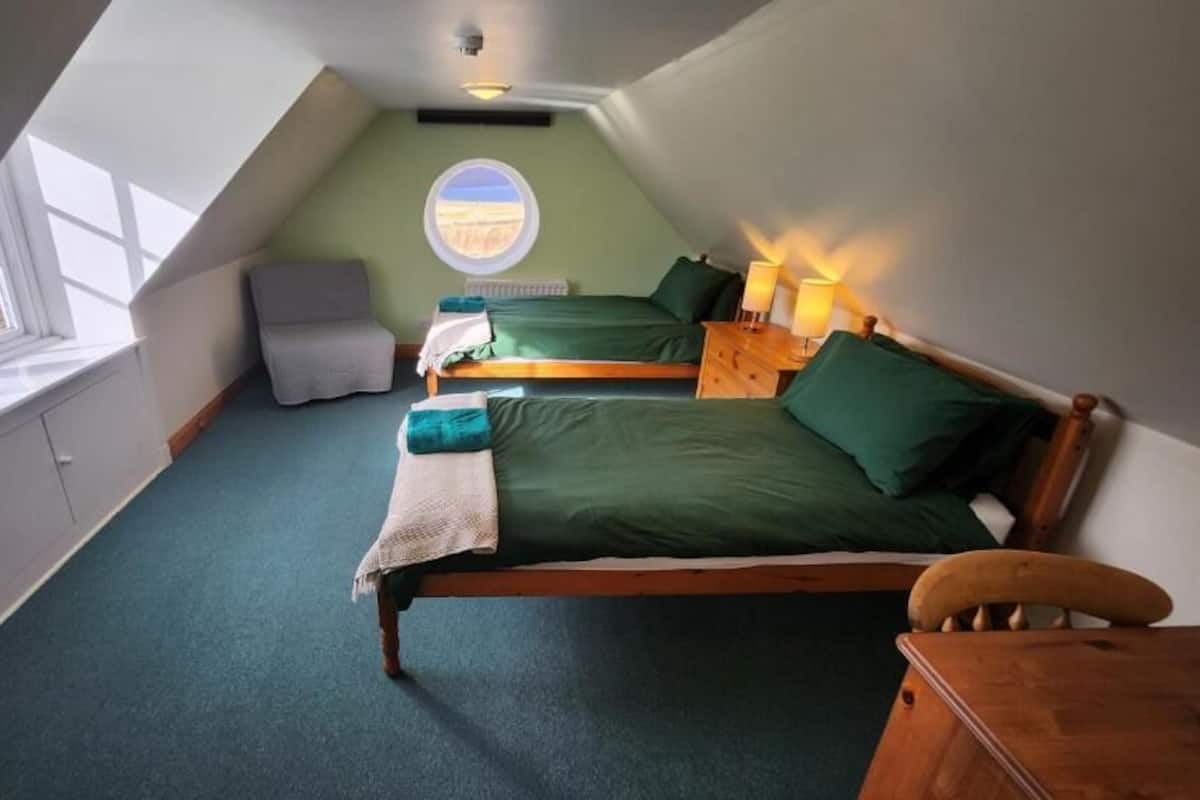 A bedroom with a sloped ceiling, two beds, a chair, a window, and a view of the outdoors.