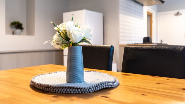 Una mesa de comedor con un florero azul con flores blancas, un mantel tejido y una silla negra.
