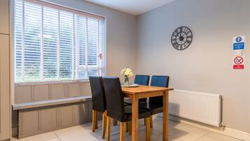 A dining area with a wooden table, four chairs, a clock, and a window with blinds.