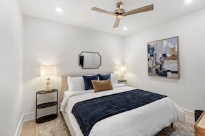 A hotel room with a bed, two bedside lamps, a ceiling fan, a painting, and a small table with a lamp.