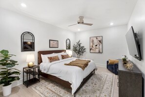 Een slaapkamer met een bed, nachtkastjes, een plant in pot, een plafondventilator en wanddecoratie.