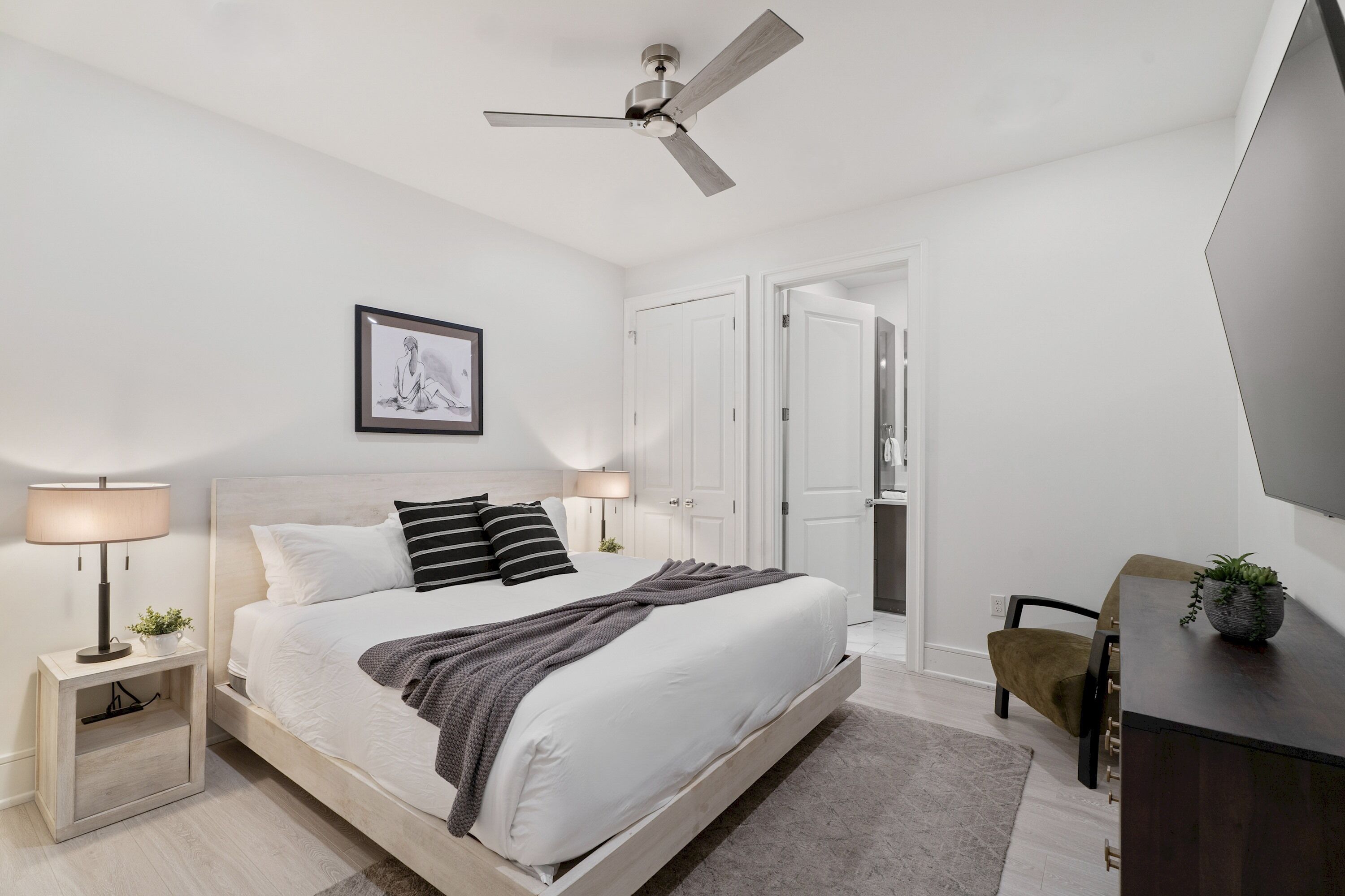 A modern bedroom with a bed, bedside tables, a ceiling fan, a framed picture, a desk with a plant, and a TV.