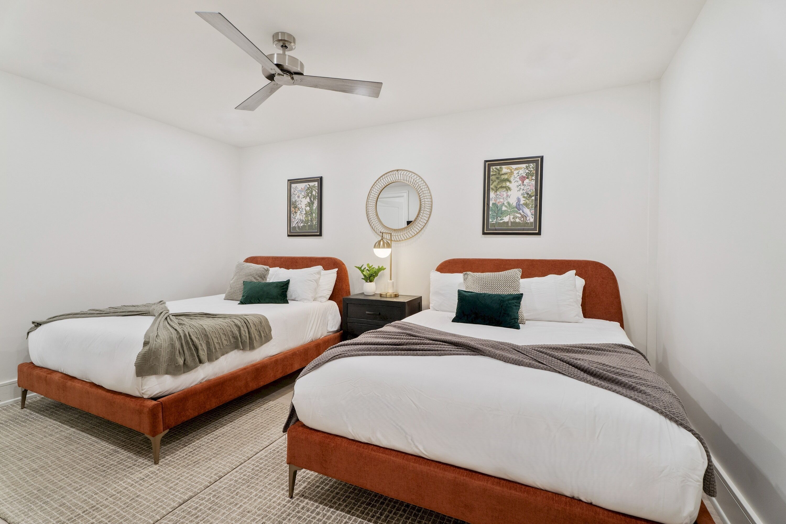 A bedroom with two beds, a ceiling fan, a nightstand with a lamp, and framed pictures on the walls.