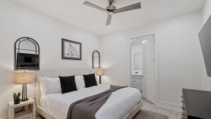 A modern bedroom with a bed, bedside lamps, a ceiling fan, a framed artwork, a mirror, and a TV.