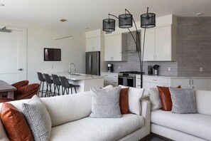 Relax in the living room with a sofa and dining area, featuring a kitchen with a refrigerator, stove, and pendant lights.