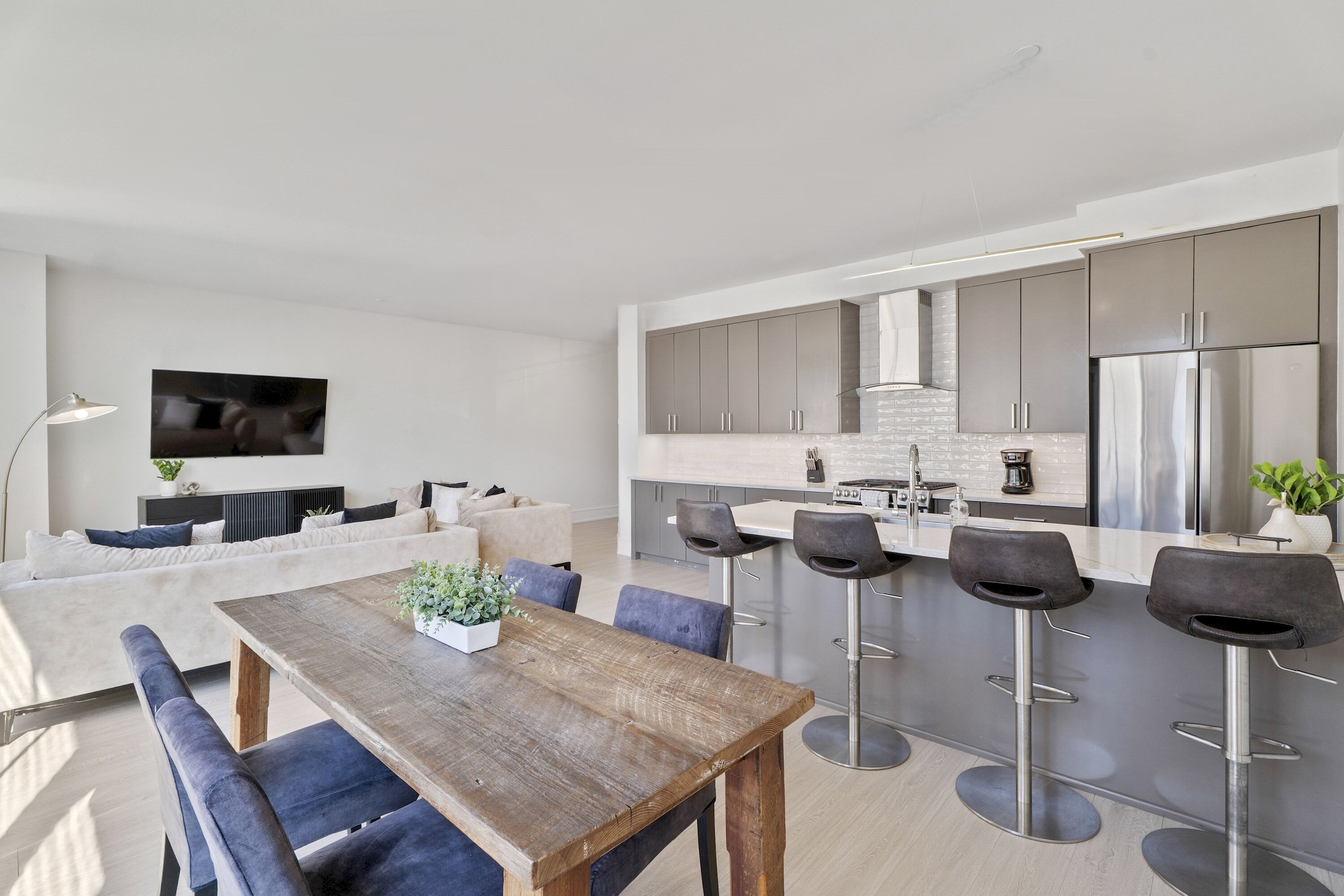 Eine moderne Küche mit Edelstahlgeräten, ein Essbereich mit Holztisch und Barhocker sowie ein Wohnbereich mit Sofa und Flachbildfernseher.