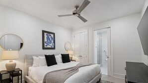 Un dormitorio con cama, mesitas de noche, ventilador de techo, una obra de arte enmarcada y una puerta que da al baño.