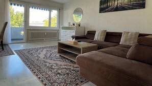 A living room with a brown sofa, a coffee table, a rug, a white cabinet, and a painting of a forest path. - Very nice apartment in the heart of Interlaken with all amenities (Matten bei Interlaken)