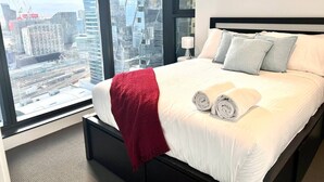 Habitación de hotel con una cama grande, una manta roja y dos almohadas. La habitación ofrece vistas de un paisaje urbano a través de amplios ventanales.