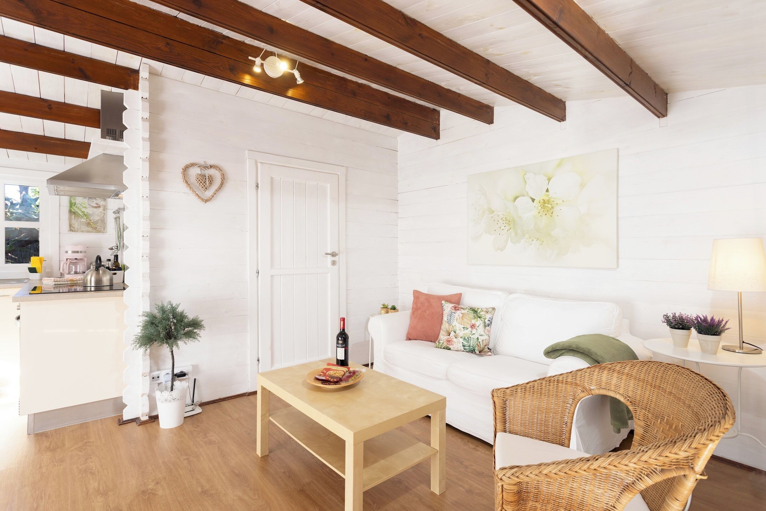 Uma sala de estar aconchegante com um sofá branco, uma mesa de centro de madeira e uma poltrona de vime. Uma pequena mesa com luminária e plantas em vasos fica no canto.