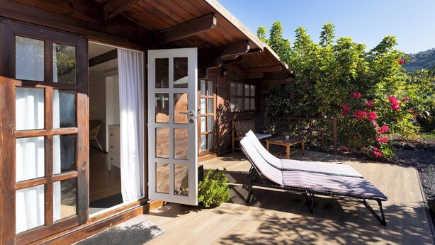 A wooden cabin with a sliding glass door, a lounge chair, and a garden with pink flowers. - Home2Book Comfy Cottage Hibiscus, Peace Retreat (La Orotava)
