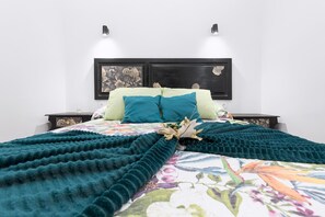 A neatly made bed with a floral bedspread and a teal blanket. There are two wall-mounted light fixtures on either side of the bed.