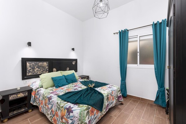 A bedroom with a floral bedspread, teal curtains, a geometric light fixture, and a nightstand with a drawer.