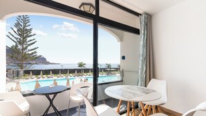 A balcony with a view of a pool and mountains, featuring white chairs and a round table. - Home2Book Cosy Studio, Pool & Sea Views Mogán (Mogán)