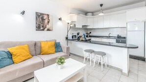 Una cocina moderna con armarios blancos, encimera gris y microondas integrado. Hay un sofá blanco con cojines amarillos y una pequeña mesa con una planta.