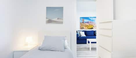A minimalist bedroom with a bed, bedside table, and a view of a living area with a blue sofa and a painting.