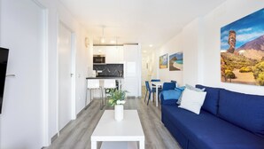 A modern living room with a blue sofa, a white coffee table, and a TV on the left. A kitchen is visible in the background with white cabinets and a dining area with blue chairs. - Home2Book Ocean Breeze Terrace Stay, Mesa del Mar (Mesa del Mar)