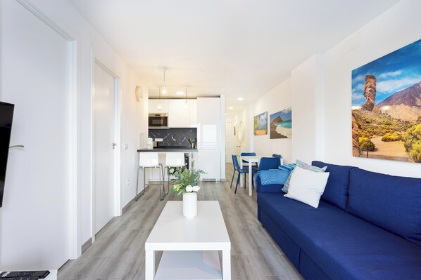 A modern living room with a blue sofa, a white coffee table, and a TV on the left. A kitchen is visible in the background with white cabinets and a dining area with blue chairs.