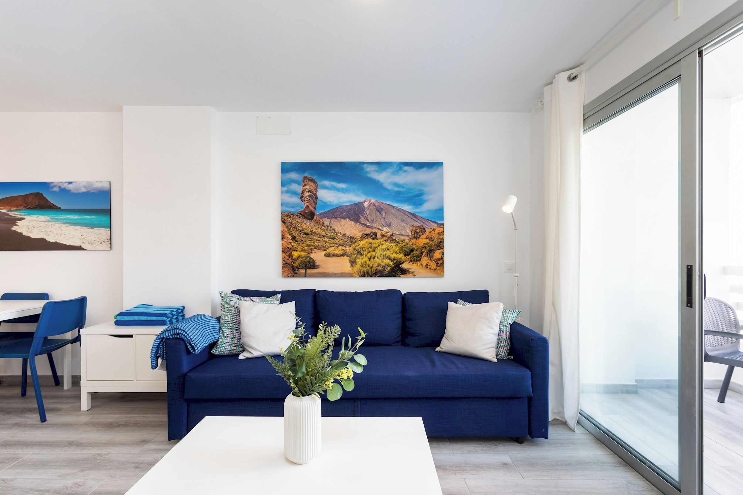 A modern living room with a blue sofa, white cushions, and a white coffee table with a plant. There is a painting of a mountain landscape on the wall.