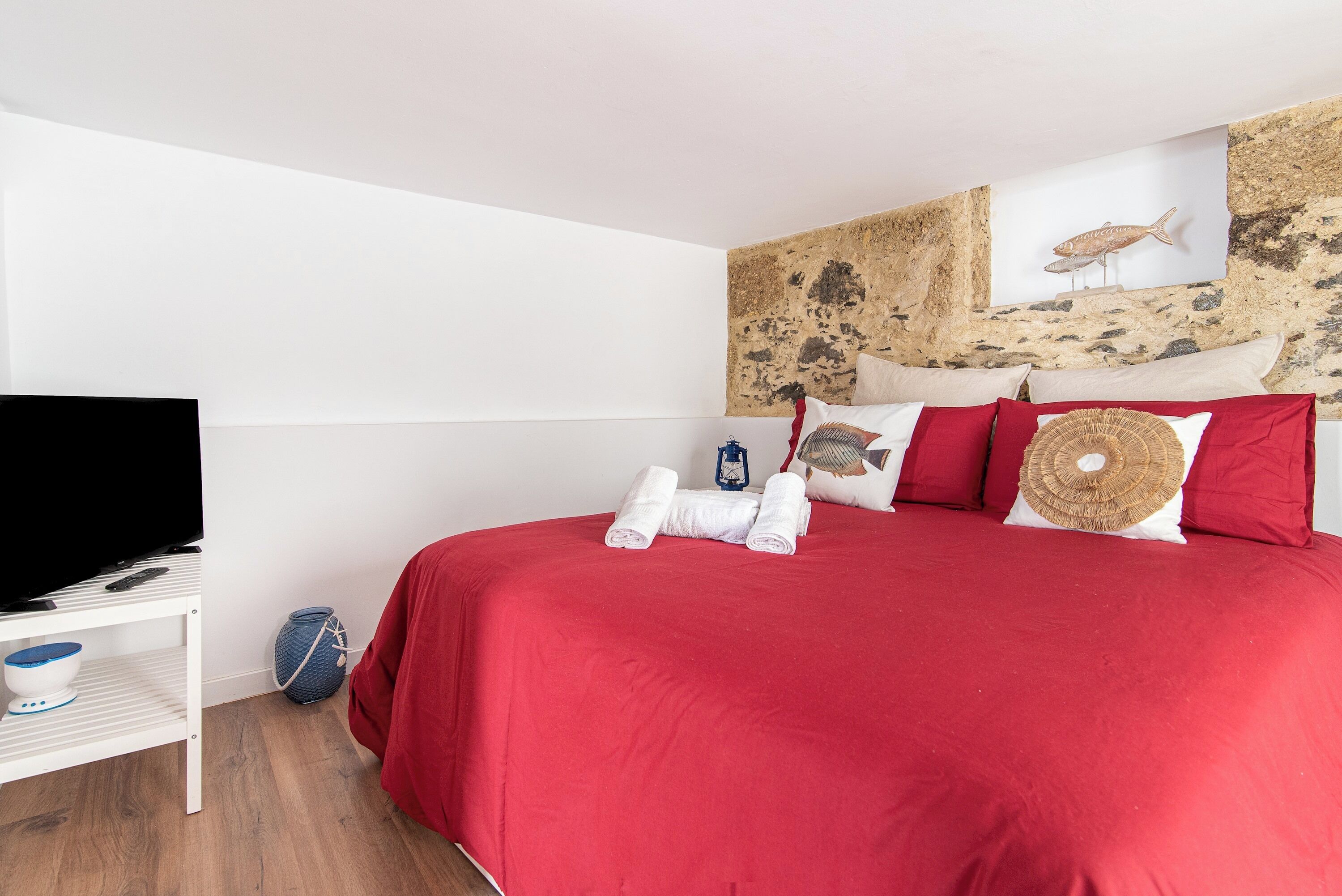 A hotel room with a large bed, a flat-screen TV, a small shelf with a bowl and a vase, and a stone wall with a fish decoration.