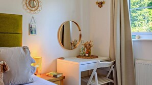 A bedroom with a bed, bedside table, lamp, mirror, and a view of greenery outside.