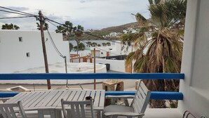 A balcony with a table and chairs, overlooking a coastal town with white buildings and palm trees. - Ornos Beachside 2 Bd Stylish House (Mykonos)