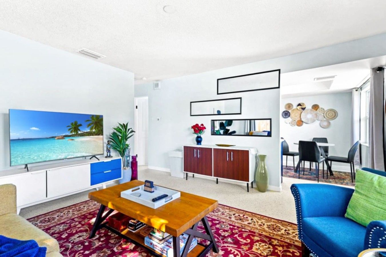 A living room with a flat-screen TV, a wooden coffee table, a blue sofa, a dining area with a white table and chairs, and a decorative wall with mirrors and plates.