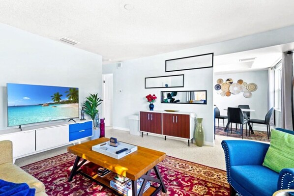 A living room with a flat-screen TV, a wooden coffee table, a blue sofa, a dining area with a white table and chairs, and a decorative wall with mirrors and plates. - Charming Villa 14 mins away from Siesta Key Beach (Sarasota)