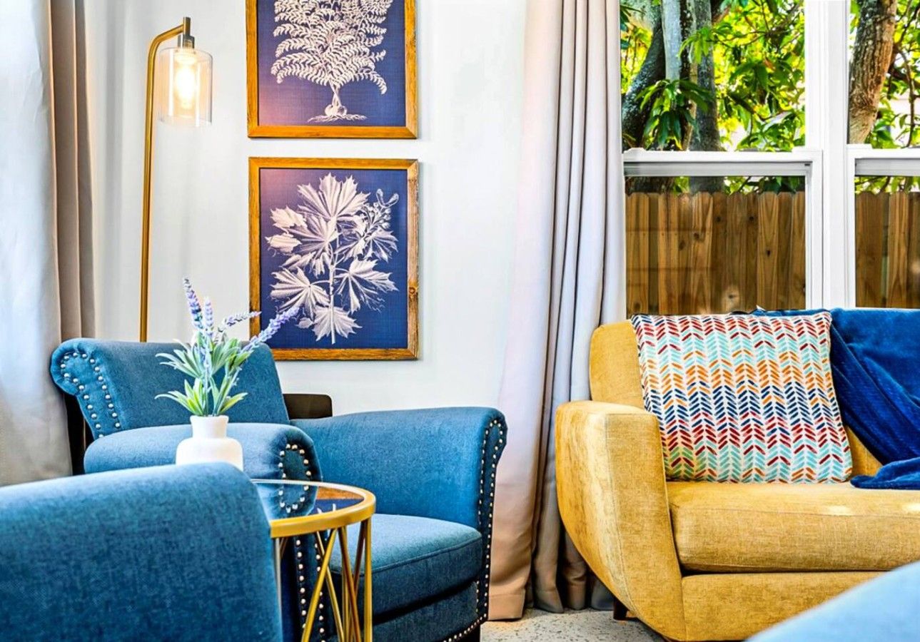A room with two sofas, one blue and one yellow, a small table with a plant, and two framed botanical prints on the wall.