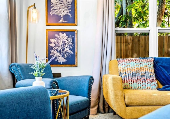 A room with two sofas, one blue and one yellow, a small table with a plant, and two framed botanical prints on the wall.