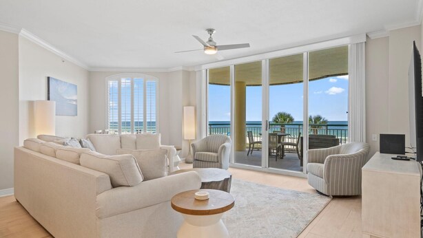A spacious living room with a large sofa, a round coffee table, and a TV stand. The room has a ceiling fan and large windows offering a view of the ocean. - Beach Colony East 1B (Pensacola)