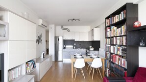 A modern kitchen with white cabinetry, a dining area with a wooden table and chairs, and a bookshelf filled with books.