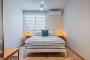 A bedroom with a bed, a ceiling fan, an air conditioner, two bedside lamps, and a window with blinds.