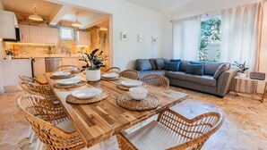 A modern living space with a wooden dining table set for six, surrounded by wicker chairs. A grey sofa and a small side table with a radio are in the background.