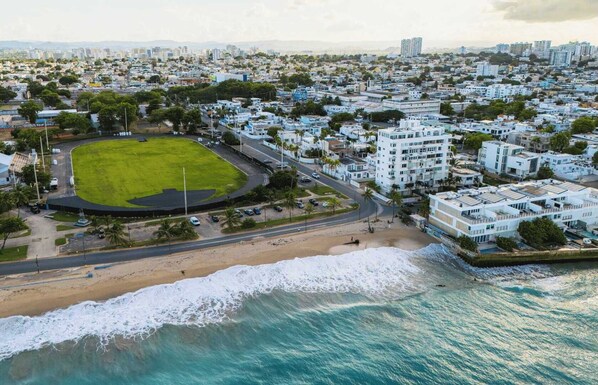 An aerial view of a coastal city with a large green sports field, a beach, and residential buildings.