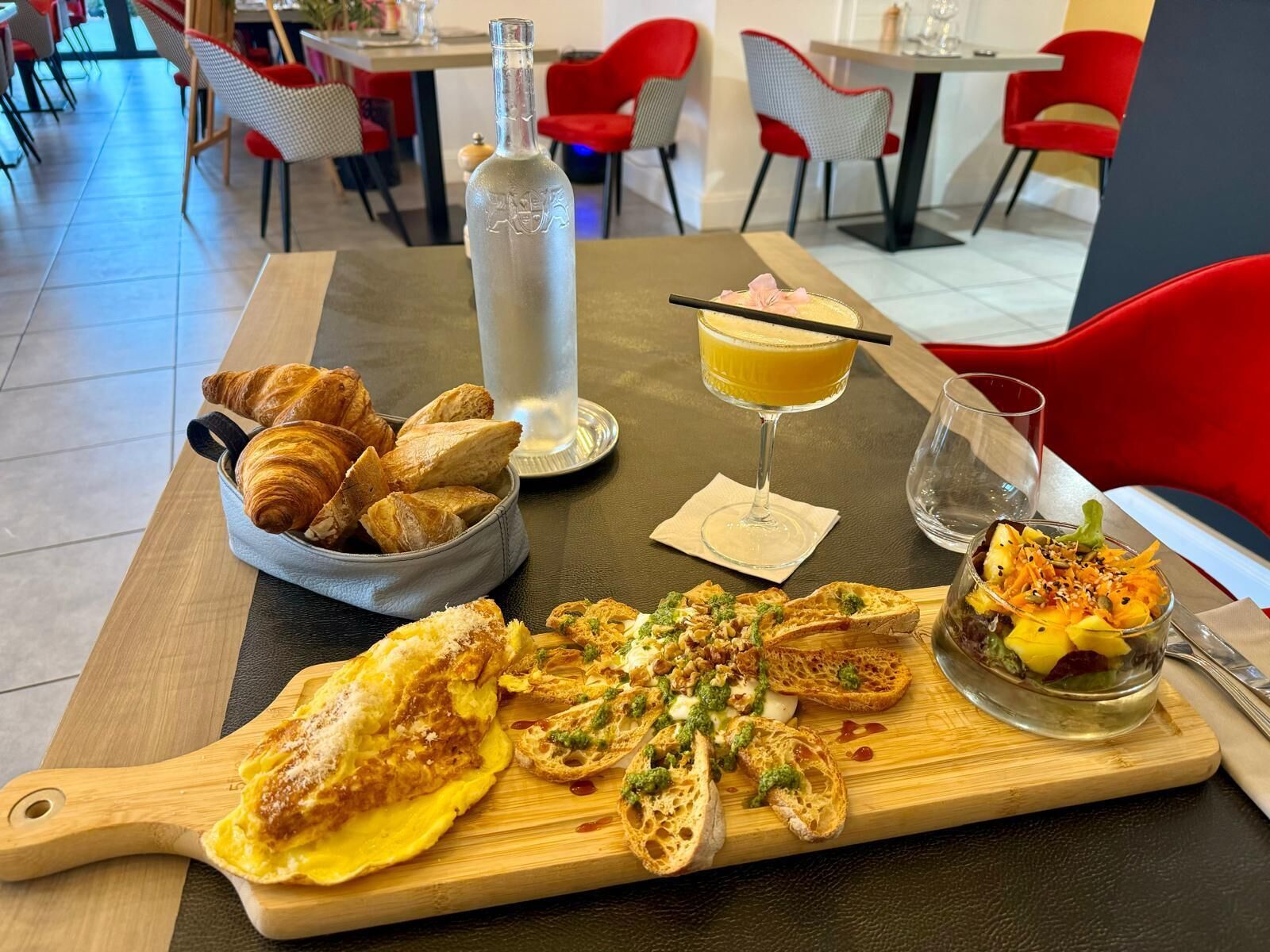 Una mesa de comedor con cruasanes, una tortilla, bruschetta, un vaso de zumo de naranja y una botella de agua.
