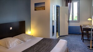 A hotel room with a bed, a desk, a chair, a radiator, and a window with curtains. - LE BON DUC (Les Billaux)