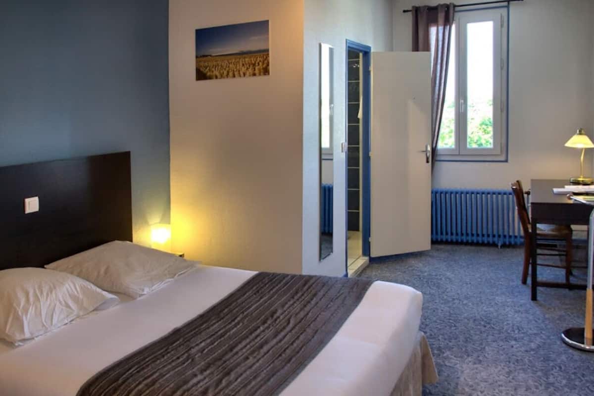 A hotel room with a bed, a desk, a chair, a radiator, and a window with curtains.