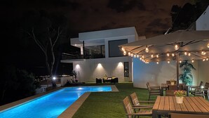 A modern outdoor pool area with artificial grass, a wooden table with chairs, and string lights.