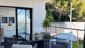 A modern outdoor patio with wicker furniture, a glass table, and a view of a lush garden and distant hills.