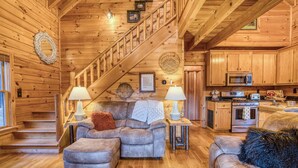 L’intérieur d’un chalet confortable, avec un escalier, un salon doté d’un canapé et d’un pouf, une cuisine équipée et des murs en bois.