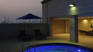 Una piscina interior con dos sillones de descanso y una sombrilla, un horizonte urbano al fondo, y un edificio moderno con iluminación cálida en su interior.