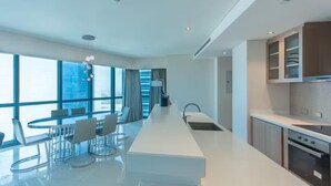 Une cuisine moderne dotée d’un îlot central, d’un coin repas avec une table ronde et des chaises, et de grandes fenêtres offrant une vue sur la ville.