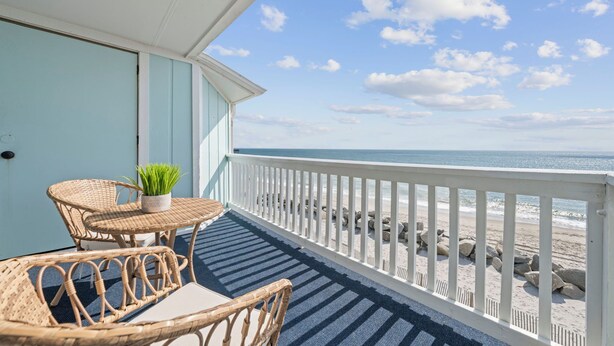 A balcony with wicker furniture and a potted plant, overlooking a beach with white railings and rocks. - A Little Shore Thing- 1 Bed Oceanfront Condo (Carolina Beach)