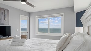 A bedroom with a large bed, a flat-screen TV, a ceiling fan, and a view of the ocean through a large window. - A Little Shore Thing- 1 Bed Oceanfront Condo (Carolina Beach)