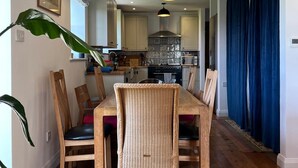 Un comedor con una mesa y sillas de madera, una cocina al fondo y una cortina azul a la derecha.