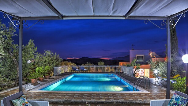 Une piscine entourée de chaises longues et d’une table où sont servis des boissons, le tout agrémenté d’un patio couvert offrant une vue sur les montagnes au crépuscule.