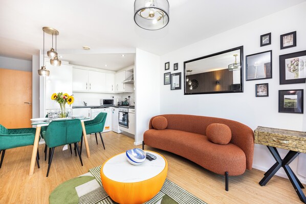 A modern living room with a sofa, a dining table with chairs, and a kitchen in the background. - Hide Out Two Bed Apartment at ExCeL (London)