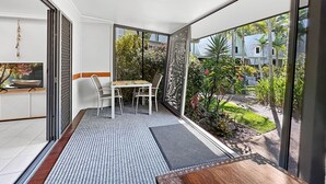 Un balcón con mesa y sillas, un televisor y vistas a un jardín y edificios.